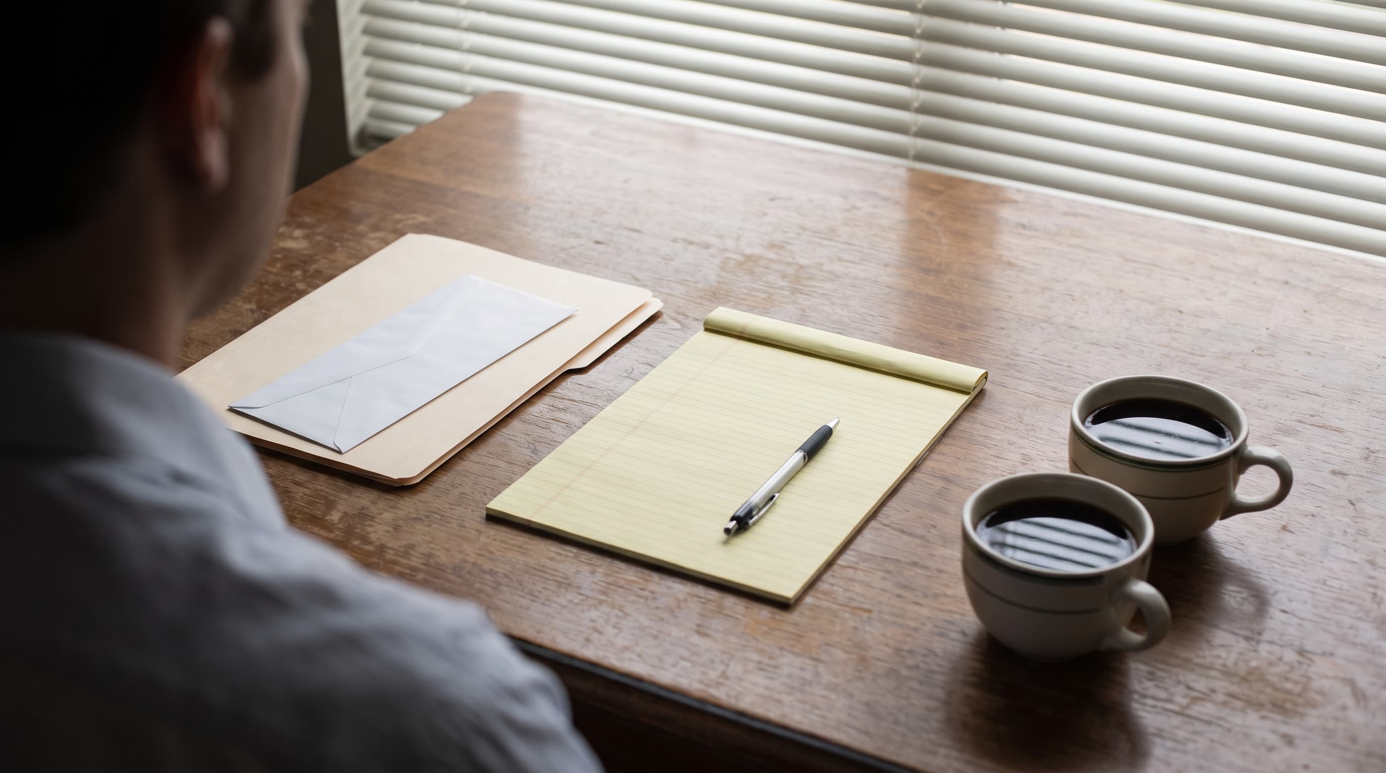 An office table seen from behind a shoulder, with two untouched coffee cups, a blank legal pad and pen, and a closed folder holding an envelope, lit by soft morning window light.