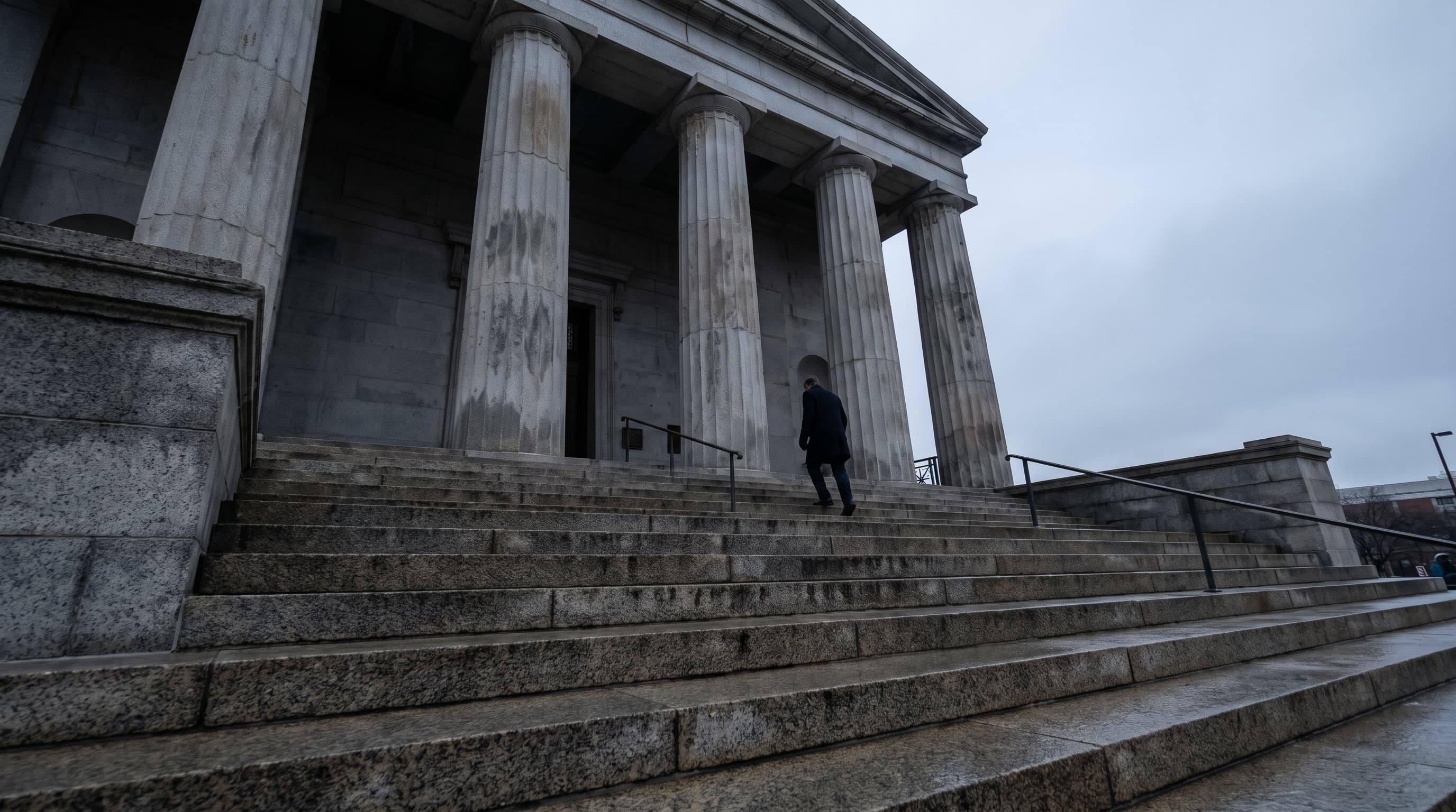 A lone figure climbs the stone steps of a courthouse under an overcast sky, columns rising above.