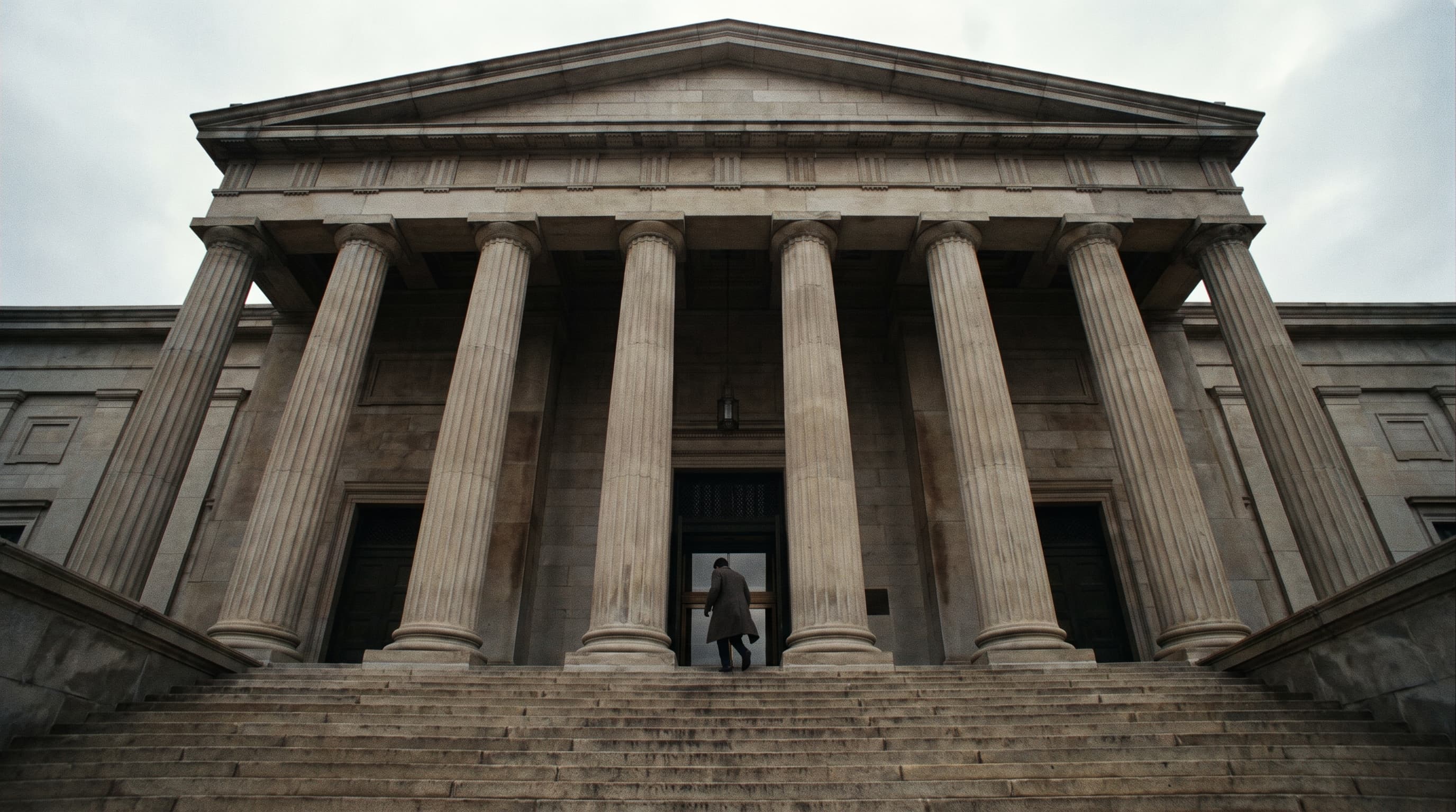 Wide view of a stone courthouse's columns and broad front steps under an overcast sky, with one small figure in a dark coat climbing toward the entrance.