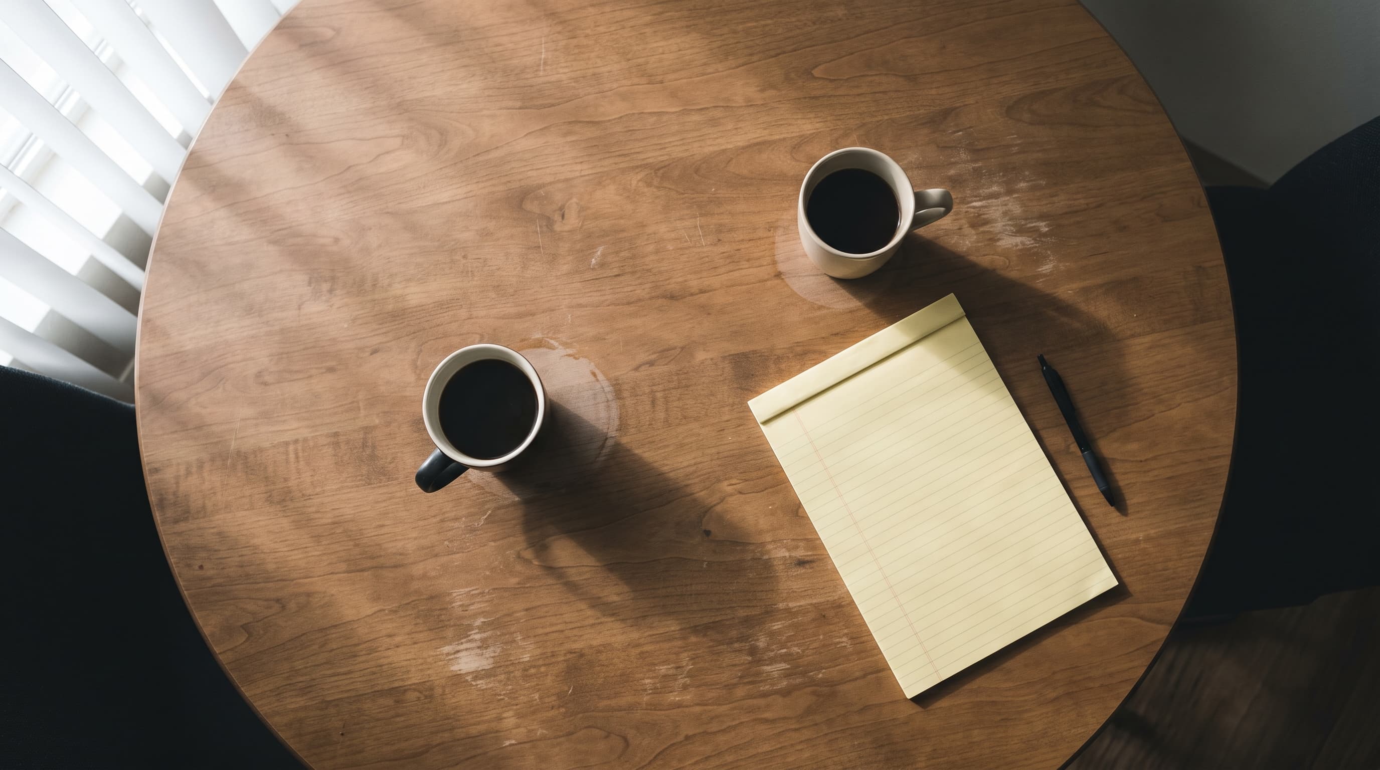 Overhead view of two coffee cups beside a closed legal pad and a pen on a wood table in soft daylight