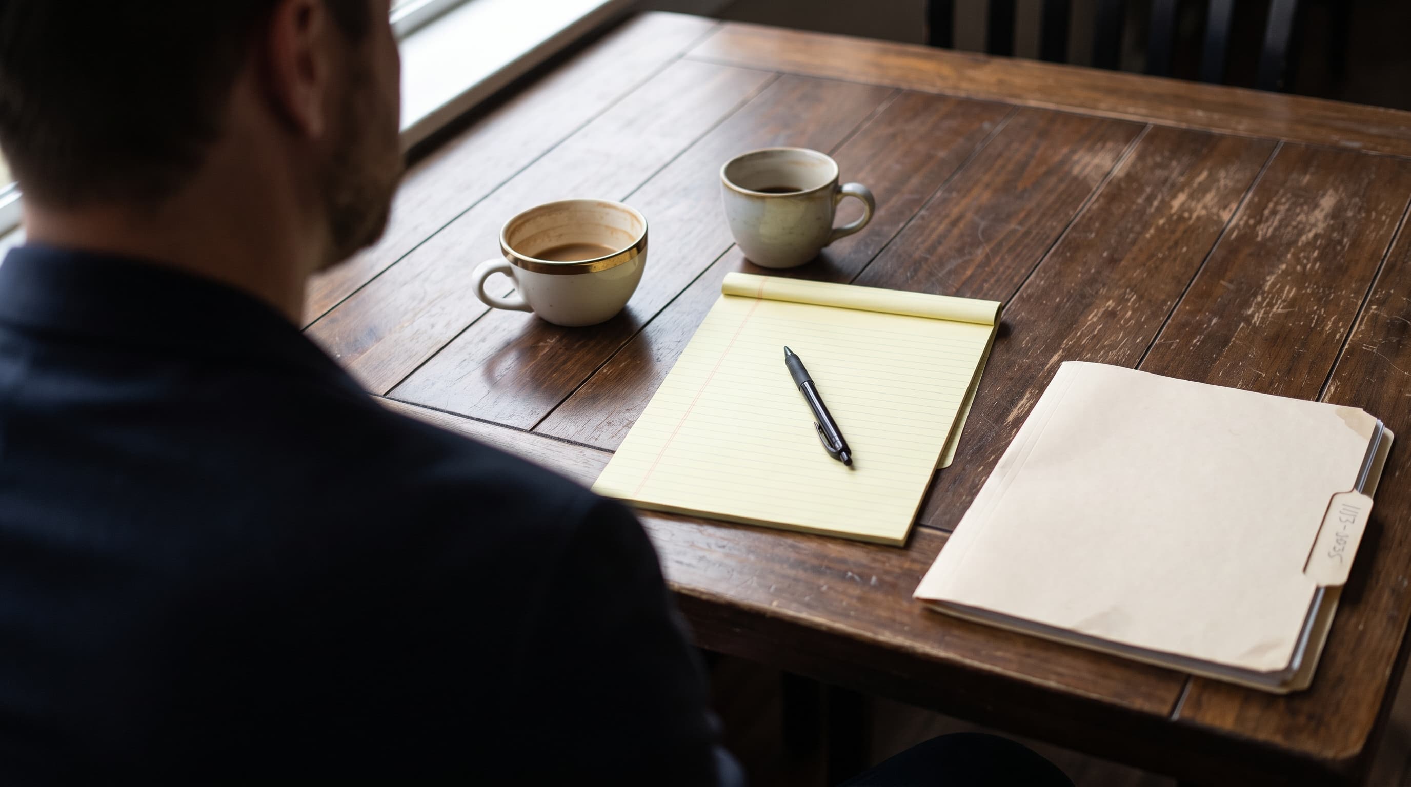 A blank legal pad and pen beside two coffee cups on a wood table, with a closed case folder nearby, seen from behind a seated person's shoulder.